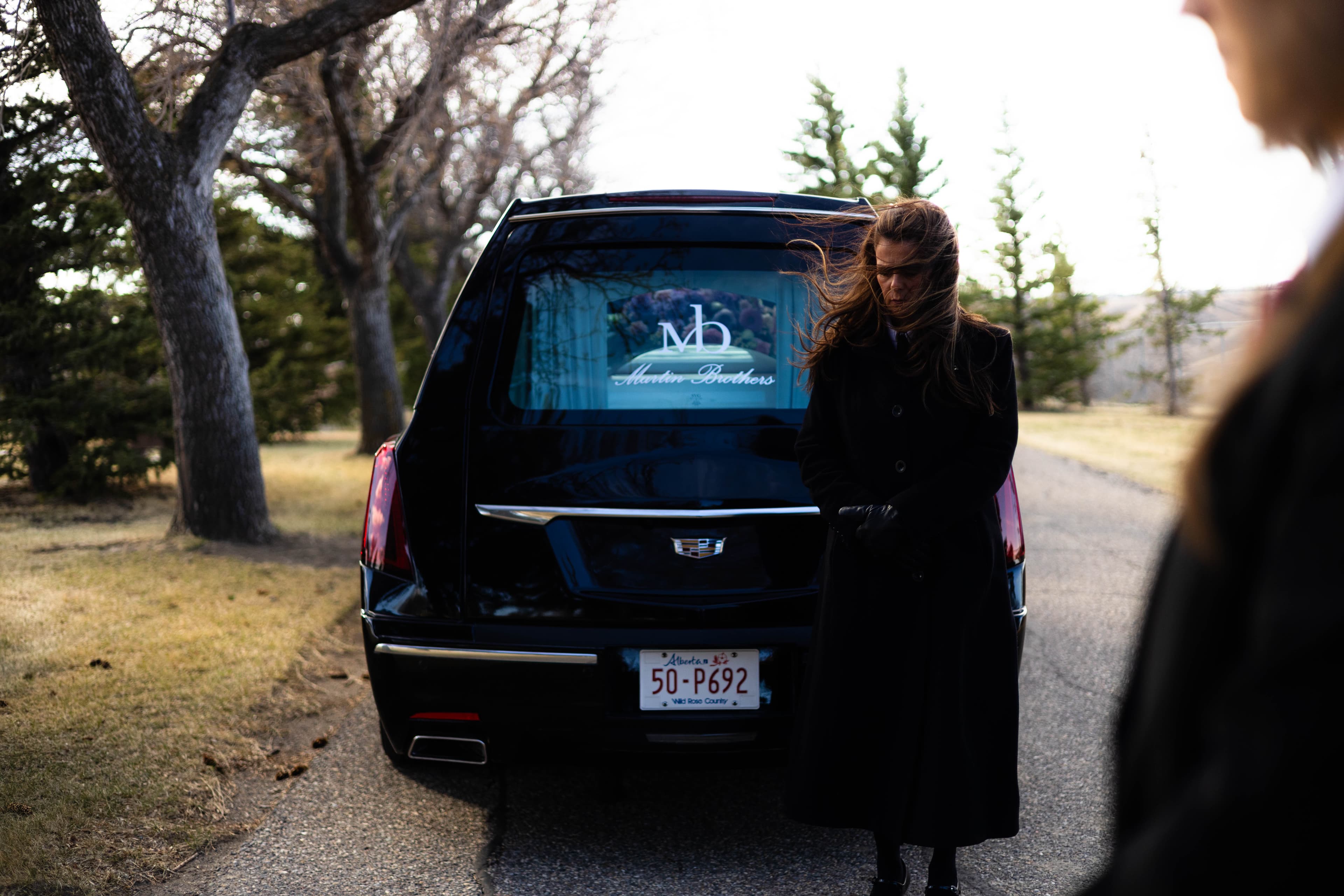 A cinematic photograph from behind a black Cadillac hearse, featuring the Martin Brothers wordmark visible through the rear window. A funeral professional in a long black coat stands in the foreground, her hair caught in the wind, against a soft-focus Southern Alberta landscape.