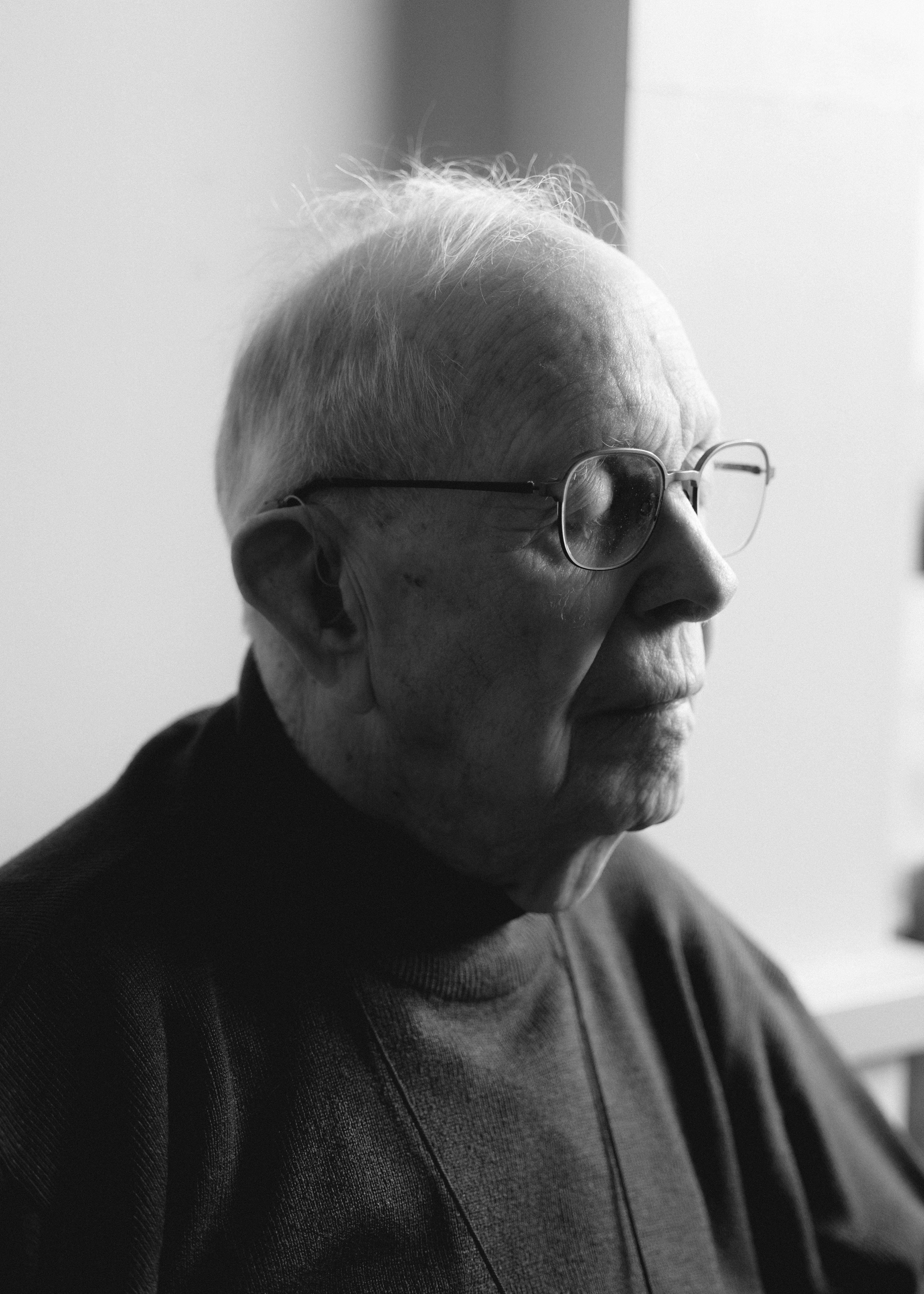 A close-up, black-and-white profile portrait of an elderly gentleman with glasses, captured in a moment of quiet contemplation. The soft, directional lighting highlights the textures of his face and the subtle drape of his dark sweater. Within this case study, this portrait exemplifies human-centered visual storytelling, using natural light and an unposed, authentic profile to communicate profound dignity, wisdom, and lived experience.