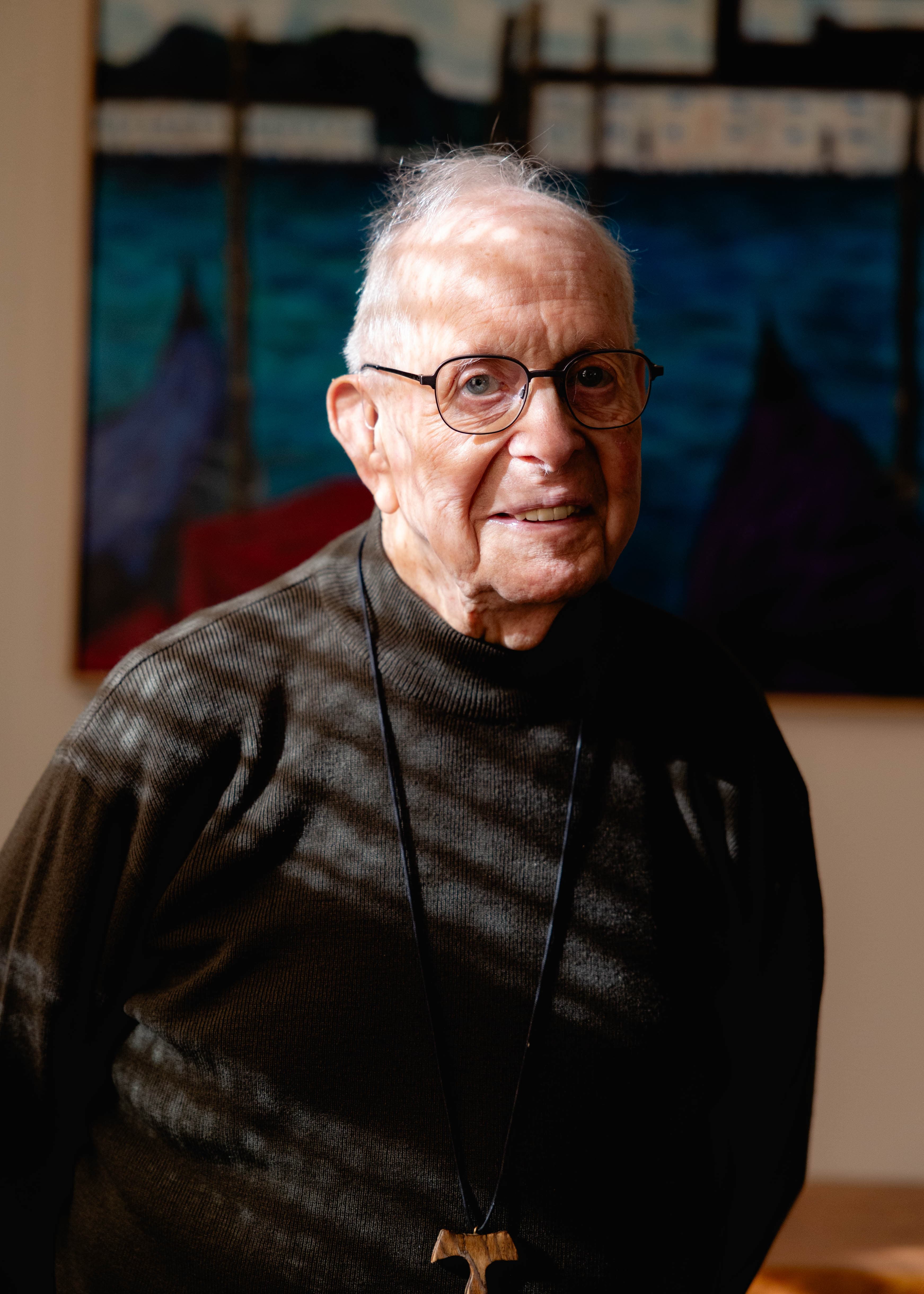 A documentary-style portrait of an elderly gentleman with glasses and a warm smile, wearing a textured dark sweater and a carved wooden cross necklace. Slatted natural light casts intentional shadows across his face and torso. In this case study, this portrait exemplifies human-centered visual storytelling, using genuine expression and intimate lighting to communicate warmth, personal history, and the profound dignity of ordinary people.