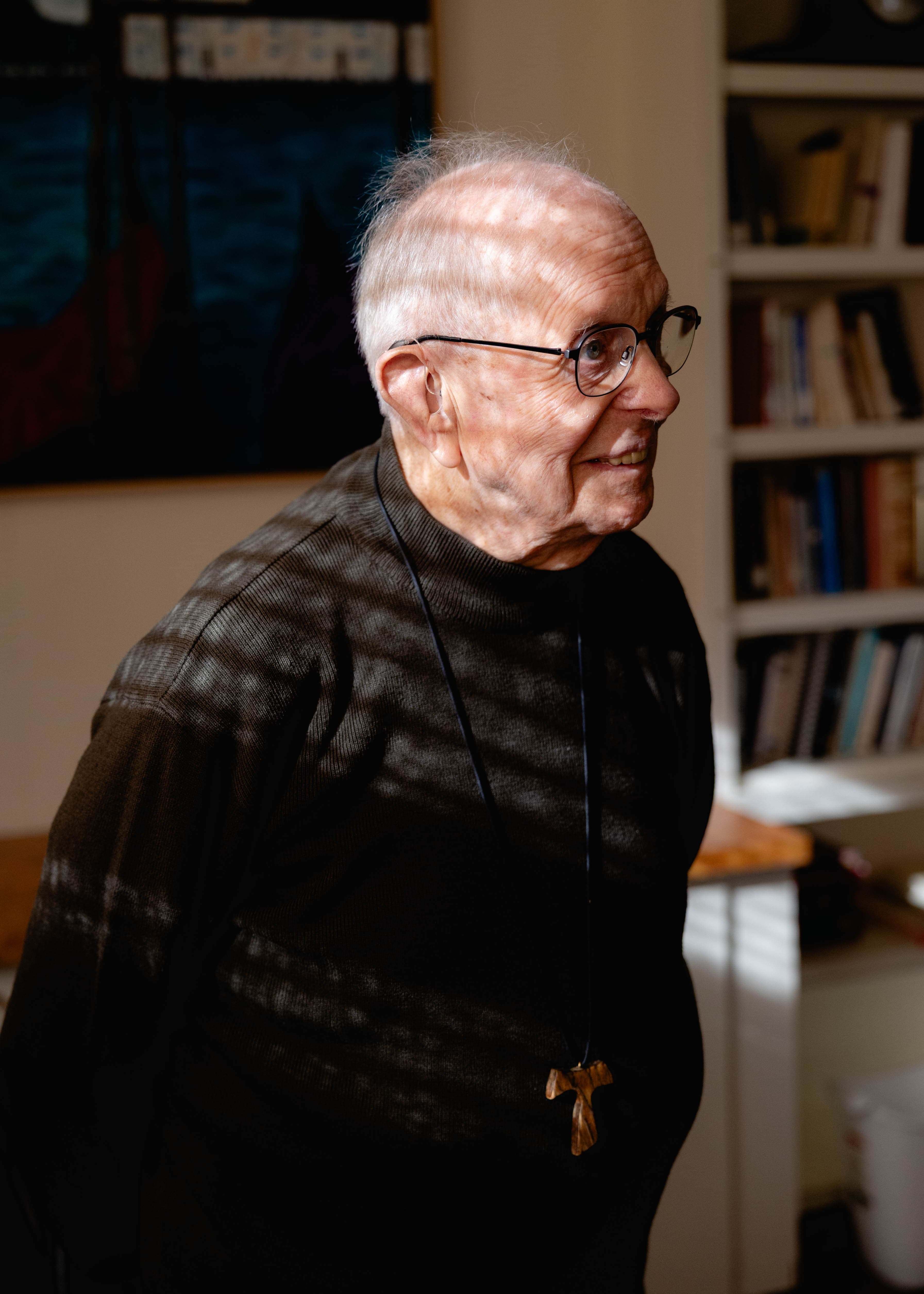 A documentary-style portrait of an elderly gentleman with glasses and a warm smile, wearing a textured dark sweater and a carved wooden cross necklace. Slatted natural light casts intentional shadows across his face and torso. In this case study, this portrait exemplifies human-centered visual storytelling, using genuine expression and intimate lighting to communicate warmth, personal history, and the profound dignity of ordinary people.