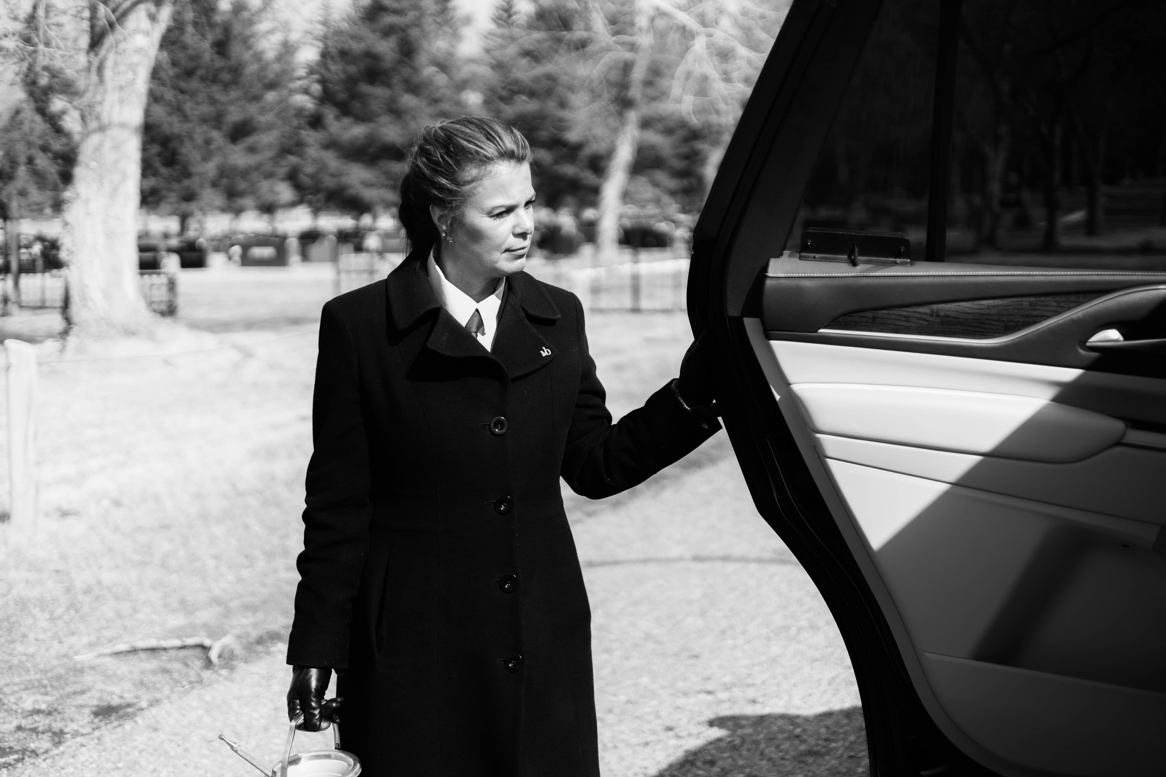 A high-contrast black and white photograph of a female funeral director in a tailored overcoat and gloves. She stands with quiet authority by an open vehicle door in a cemetery, representing the meticulous care and stewardship of the Martin Brothers brand.