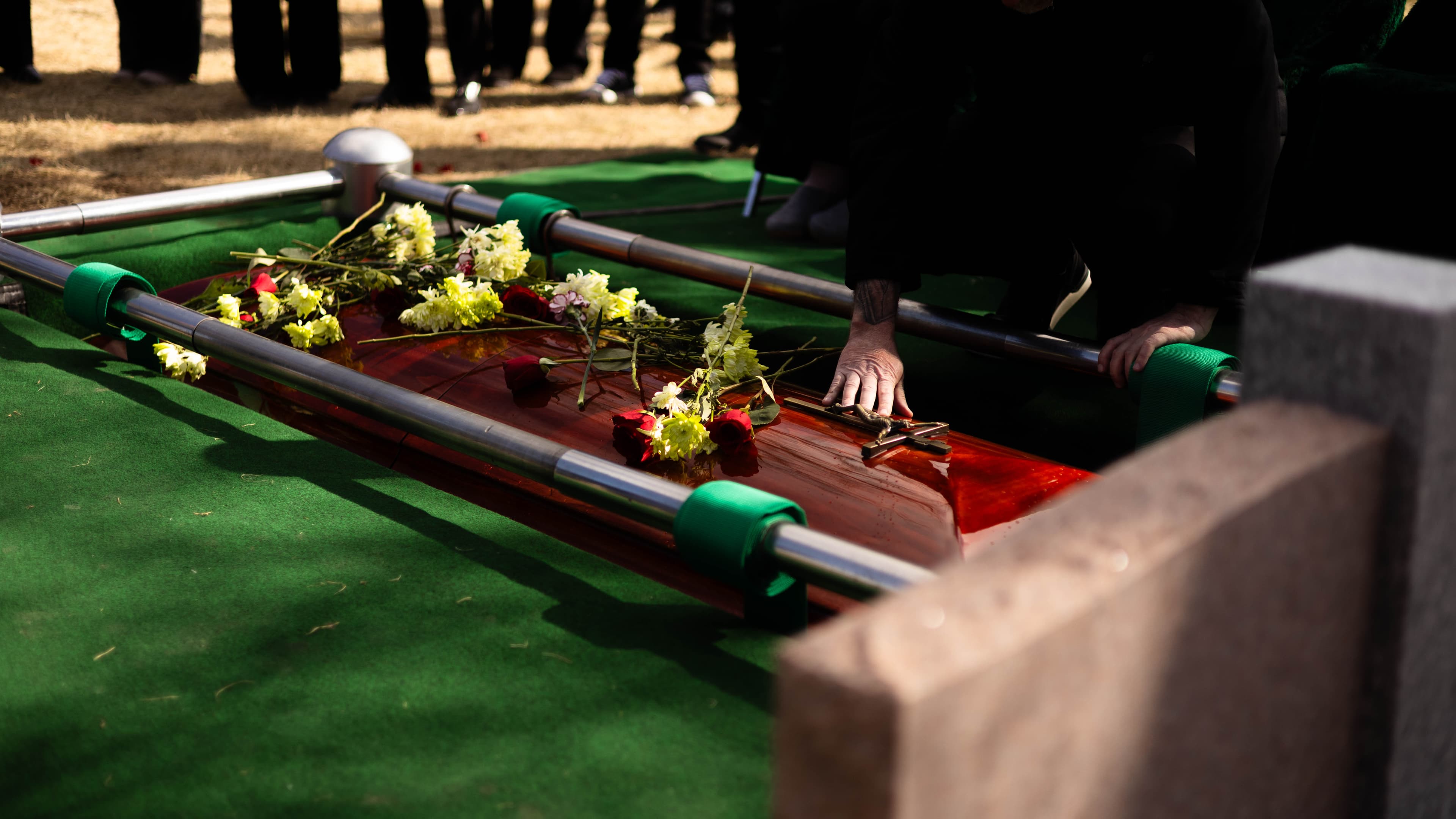 A close-up cinematic photograph of a hand resting with quiet reverence on a polished wood casket. Scattered flowers lie atop the surface, framed by the technical infrastructure of the graveside service and a blurred foreground of a granite headstone.